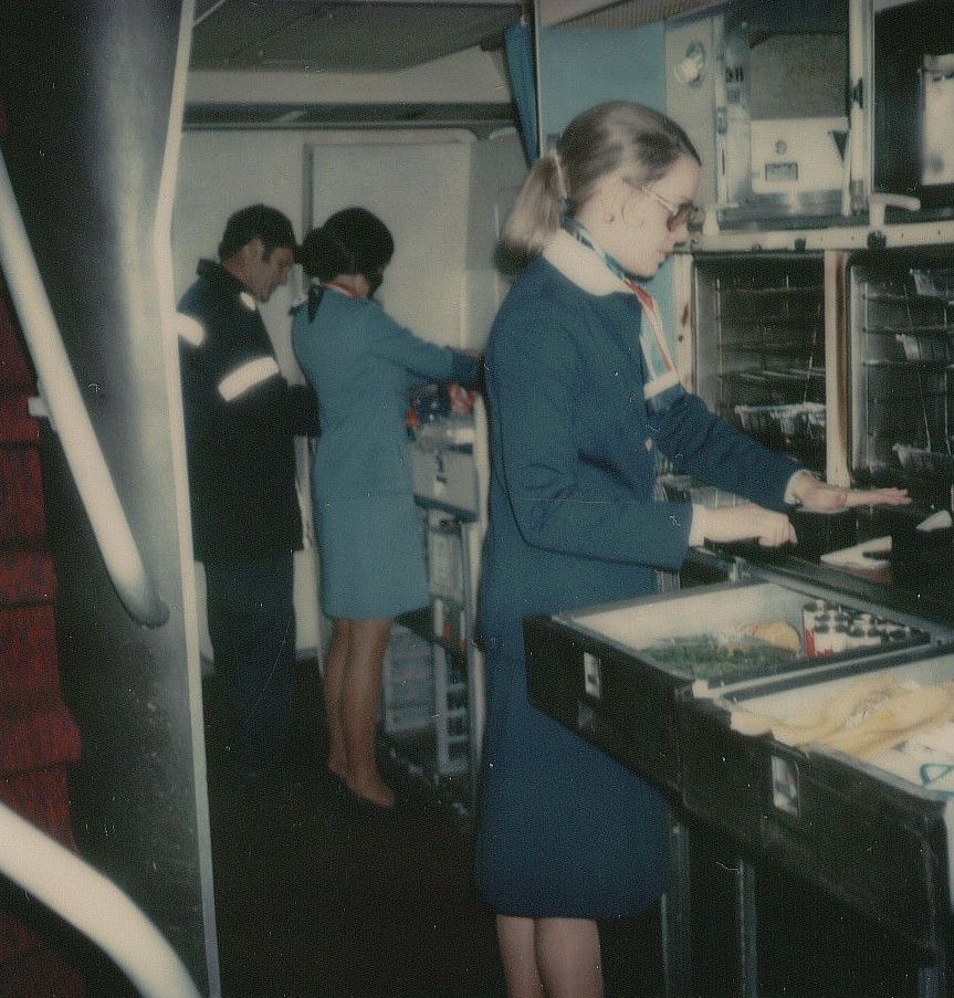 Dec 1976  A  flight attendant in navy blue prepares the first class galley of a 747 prior to customer boarding.  In  the background the  flight attendant in the light blue uniform demonstrates a broken galley shelf to a mechanic.